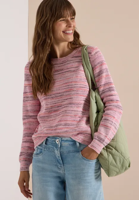 Femme souriante vêtue d’un pull rose à rayures, jean bleu et sac matelassé vert clair sur fond clair.
