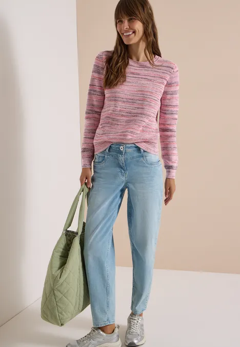 Femme avec pull rose, jean bleu clair et baskets, tenant un grand sac matelassé vert et souriant.