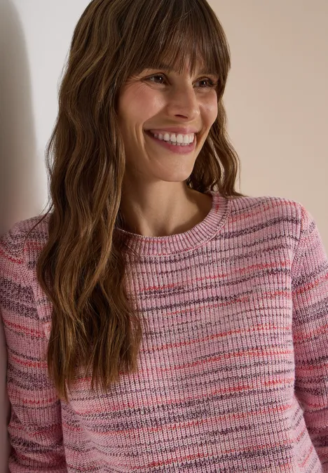 Une femme souriante aux longs cheveux bruns ondulés porte un pull rose à rayures et s’appuie contre un mur.