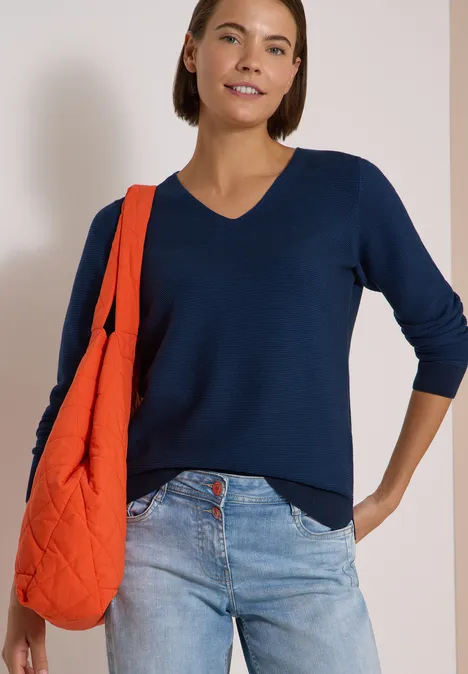 Frau in dunkelblauem V-Pullover, hellen Jeans und orangener Schultertasche vor hellem Hintergrund.
