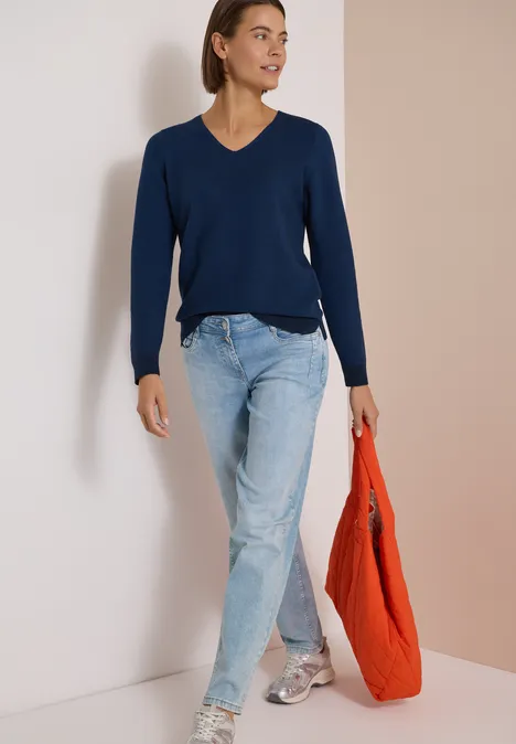 Frau in dunkelblauem V-Pullover, hellen Jeans und Sneakern, die eine orangefarbene Tasche trägt.
