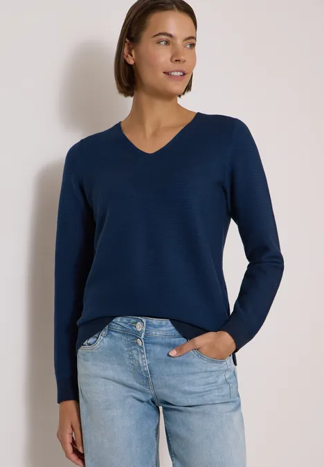 Frau trägt dunkelblauen V-Ausschnitt-Pullover und helle Jeans, lächelt vor neutralem Hintergrund.