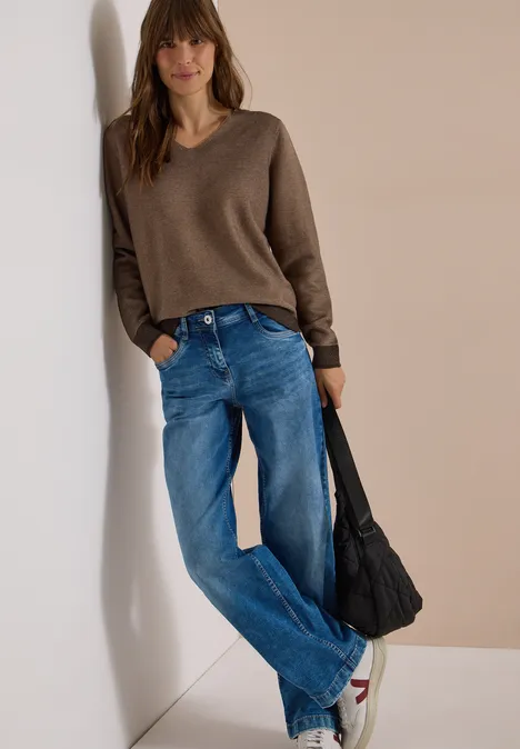 Frau mit braunem V-Pullover, blauen Jeans und schwarzer Tasche lehnt entspannt an der Wand.