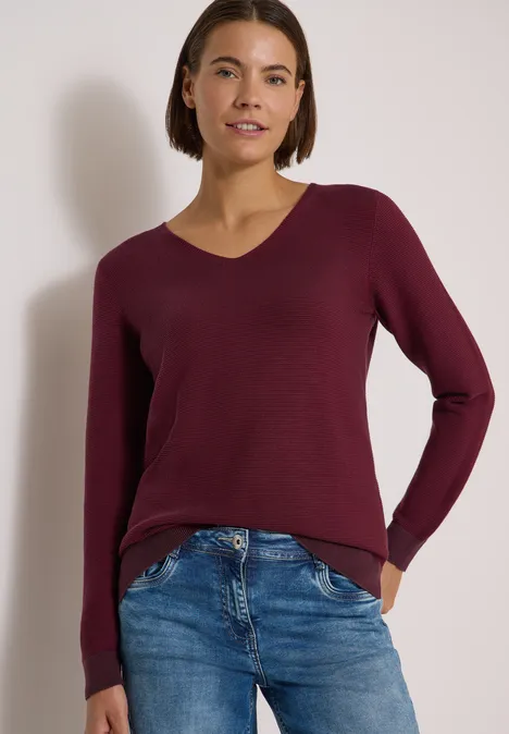 Eine Frau in burgunderrotem Pullover mit V-Ausschnitt und blauen Jeans vor schlichtem Hintergrund.
