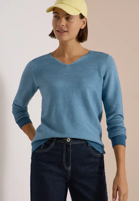 Frau mit gelber Baseballkappe, blauem V-Pullover und dunklen Jeans.