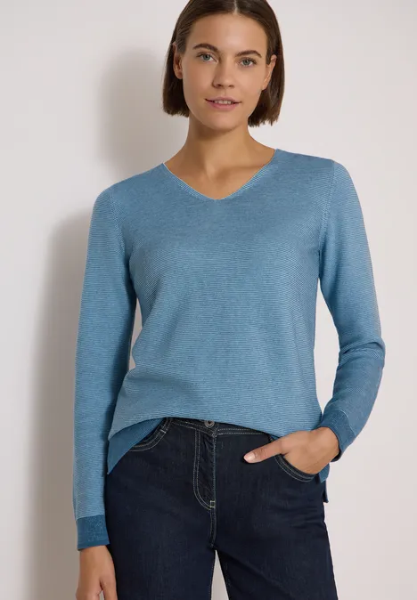 Frau im blauen V-Pullover und Jeans vor weißem Hintergrund, eine Hand in der Hosentasche.