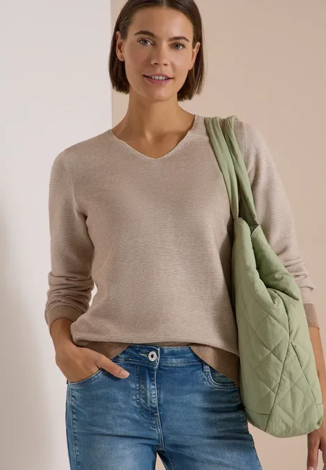 Frau in beigem Pullover und blauen Jeans mit grünem, gestepptem Shopper, lächelnd.