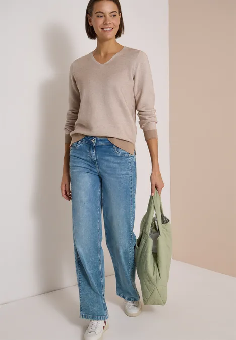 Frau mit beigem V-Pullover, weiten Jeans, weißen Sneakers und grüner Tasche vor hellem Hintergrund.