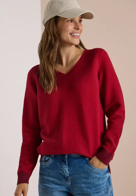 Frau in rotem V-Pullover, Jeans und Baseballcap lächelt seitlich.