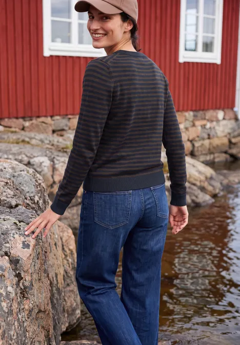 Vrouw in jeans, gestreepte longsleeve en pet, glimlachend op rotsen voor een rood houten huis aan het water.