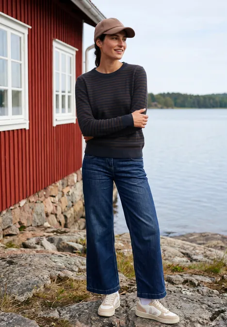 Een vrouw met gestreepte trui, wijde jeans en pet staat op een rotsachtige oever bij een rood huis aan het meer.