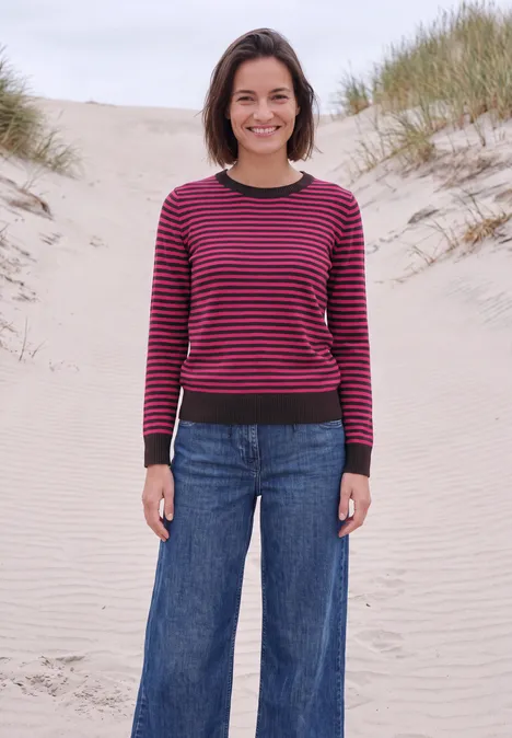Een vrouw in spijkerbroek en gestreepte trui staat op een duin bij de zee.