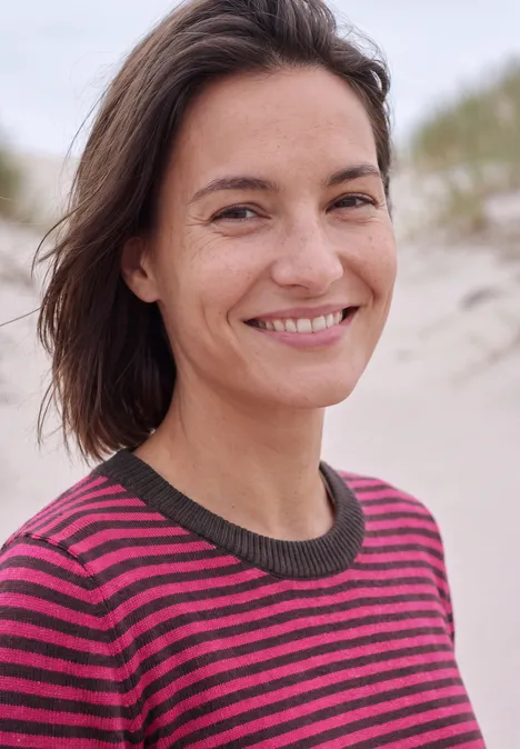 Een vrouw met bruin haar in een gestreepte trui lacht blij op het strand.