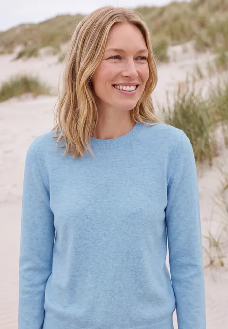 Lächelnde Frau in hellblauem Pullover steht an einem Dünenstrand.