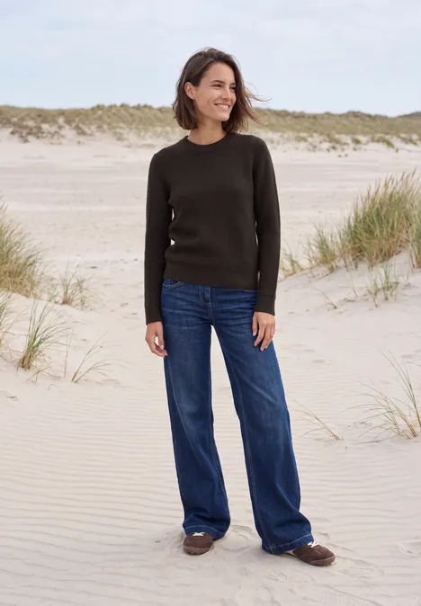 Vrouw met trui en jeans, glimlachend, staand op een zandstrand.