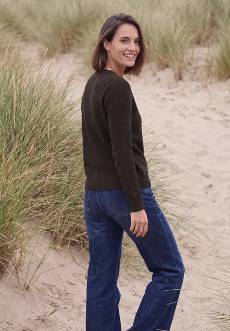 Vrouw in groene trui en spijkerbroek staat in duinen, kijkt met een glimlach over haar schouder.