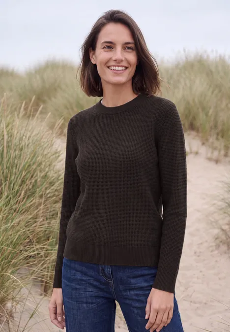 Een glimlachende vrouw staat in duinen voor hoog gras en draagt een spijkerbroek en een donker truitje.