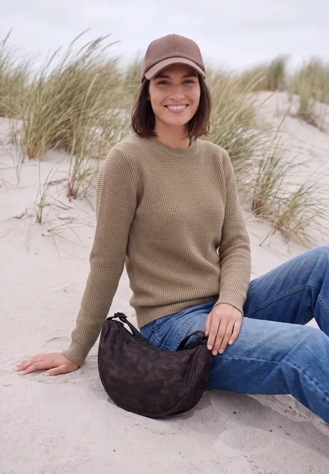 Jonge vrouw zit op een zandduin, draagt pet, trui, jeans en tas, met helmgras op de achtergrond.