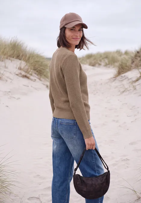 Vrouw in jeans, bruin trui en pet, met handtas bij een zandduin.