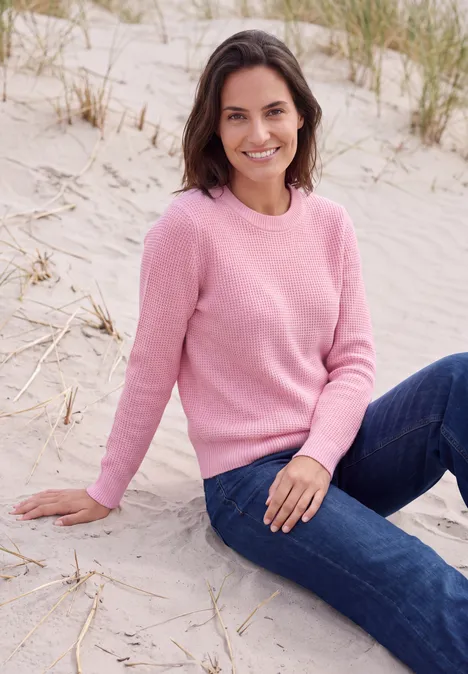 Een glimlachende vrouw in een roze trui en spijkerbroek zit ontspannen op het strandzand.