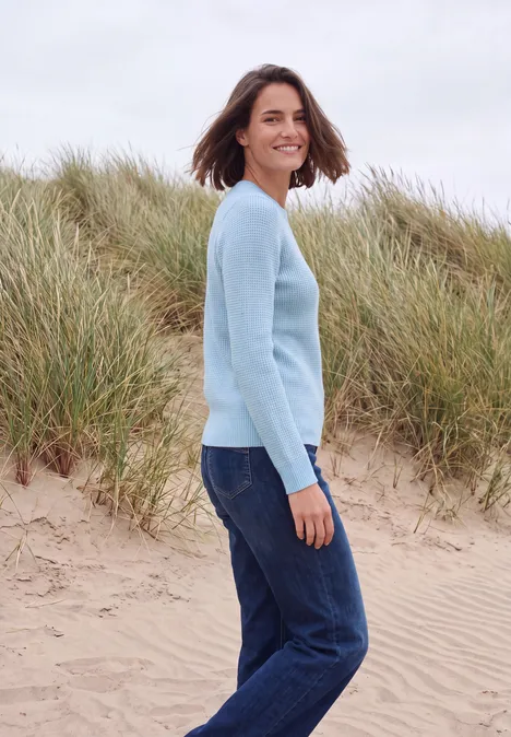 Een vrouw staat lachend op een duinenstrand, met een lichtblauwe trui en een spijkerbroek.
