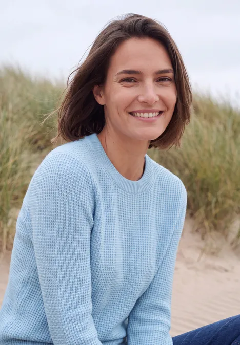 Een vrouw in een lichtblauwe trui zit glimlachend voor grasduinen op het strand.