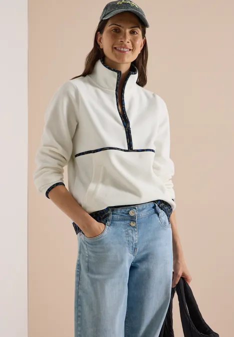 Lachende vrouw draagt een witte fleece met rits, jeans, pet en houdt een tas vast.
