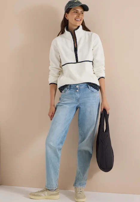 Vrouw in witte fleece met rits, lichtblauwe jeans, pet en zwarte tas.
