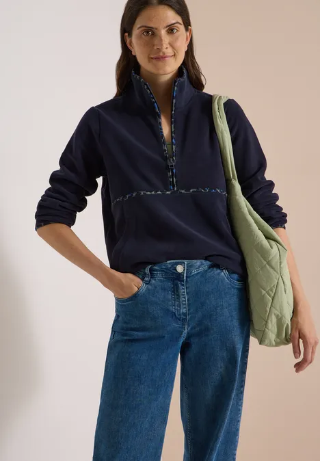 Vrouw in marineblauw fleece shirt met rits, blauwe spijkerbroek en groene schoudertas, recht vooruit kijkend.