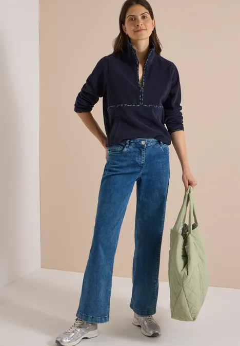 Vrouw in donkerblau trui en jeans, draagt een lichtgroene tas van stof en glimlacht ontspannen.