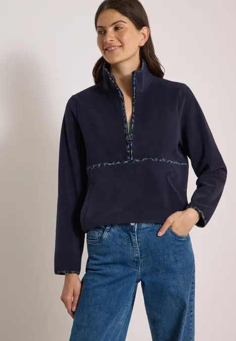 Vrouw in donkerblau fleecevest met kleurrijke bies langs ritssluiting en mouwboorden, blauwe spijkerbroek.