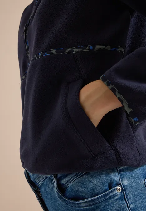 Persoon draagt donkerblauw fleecejack, hand in zak en blauwe spijkerbroek.