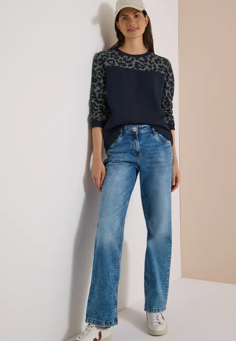Junge Frau in weiter Jeans, gemustertem Pullover, weißen Sneakern und Cap steht vor hellem Hintergrund.