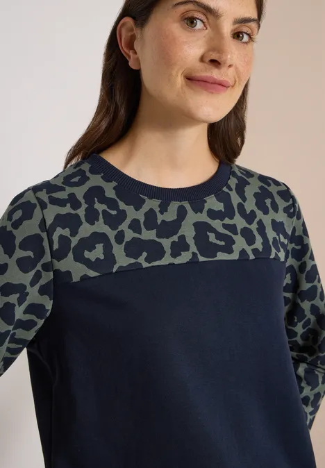 Frau im dunkelblauen Pullover mit grün-schwarzem Leopardenmuster oben und an den Ärmeln.