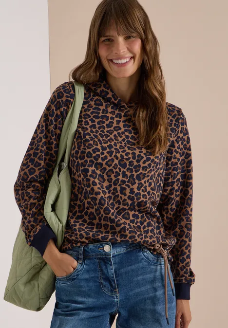 Femme souriante en sweat léopard, jean et sac vert clair.