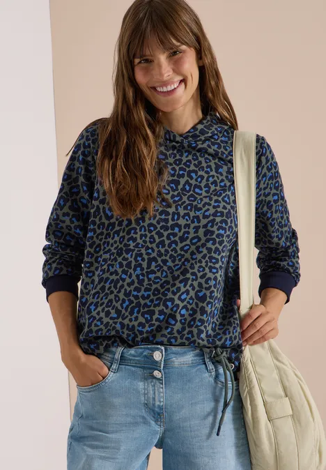 Sweatshirt mit Kapuze und Leo-Muster dried olive green Junge Frau in blauer Leo-Hoodie, Jeans und beiger Tasche, die lächelnd posiert.