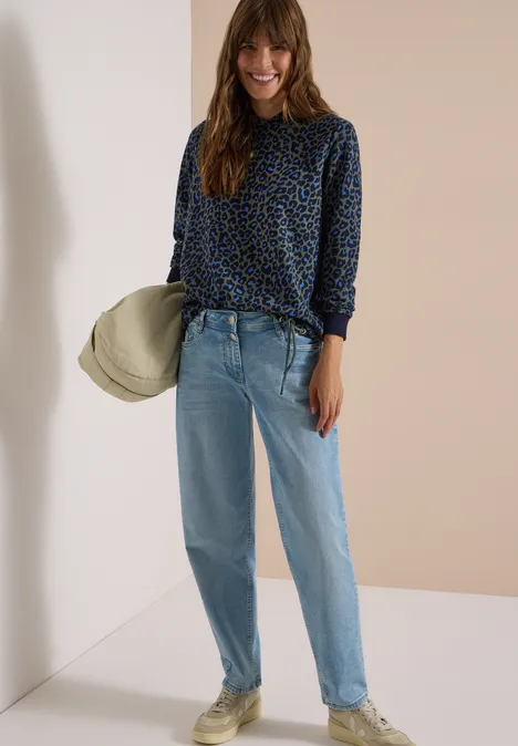 Sweatshirt mit Kapuze und Leo-Muster dried olive green Frau mit gemusterter Bluse, hellen Jeans und Hut in neutraler Umgebung.