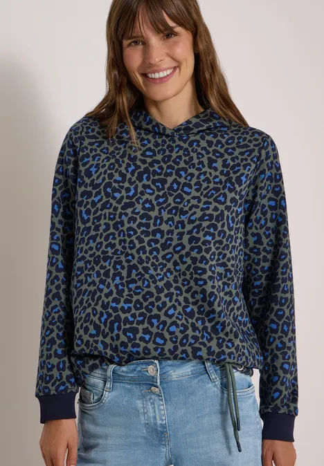 Sweatshirt mit Kapuze und Leo-Muster dried olive green Frau in blau-schwarzer Leo-Kapuzenjacke und Jeans, lächelnd vor hellem Hintergrund.