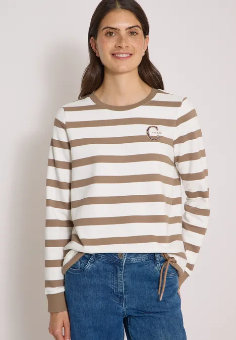 Eine lächelnde Frau trägt einen beige-weiß gestreiften Langarmpullover und blaue Jeans.