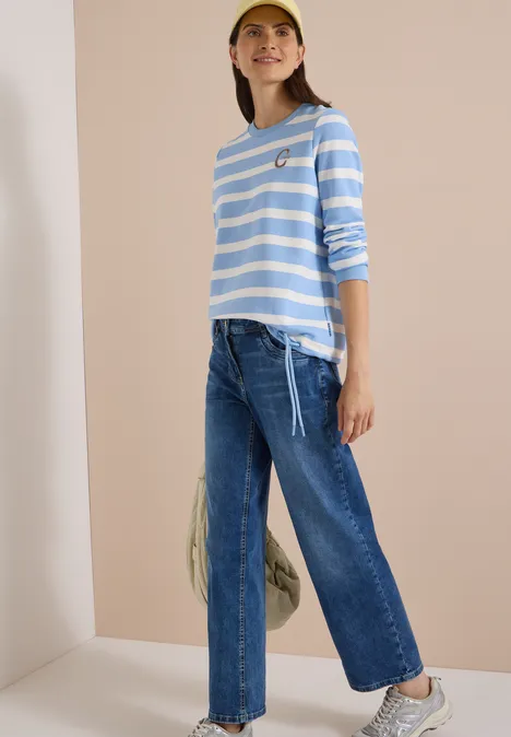 Frau steht vor hell-beigem Hintergrund, trägt blau-weiß gestreiftes Langarmshirt, weite Jeans, Sneaker und Kappe.
