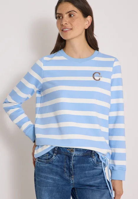 Frau trägt hellblau-weiß gestreiftes Langarmshirt mit Tunnelzug am Saum und Jeans.