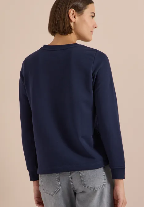 Frau von hinten, trägt dunkelblauen Pullover und graue Jeans, kurze Haare, neutraler Hintergrund.