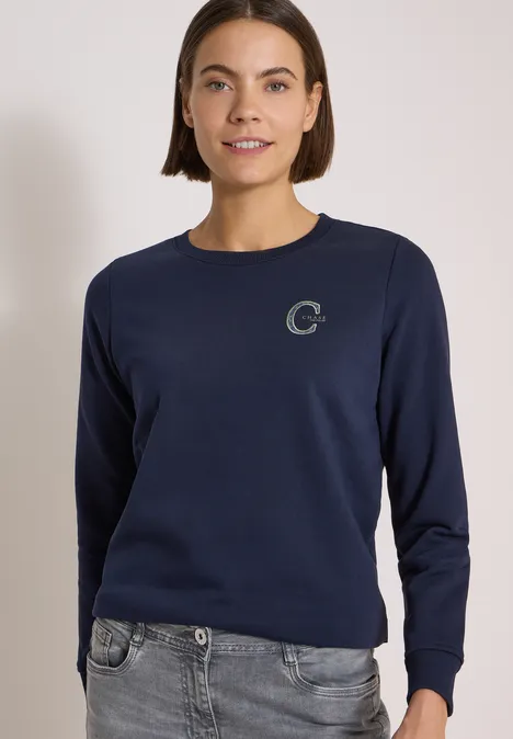 Frau trägt dunkelblaues Sweatshirt mit rundem Ausschnitt und kleinem 'C'-Logo, kombiniert mit grauer Jeans.