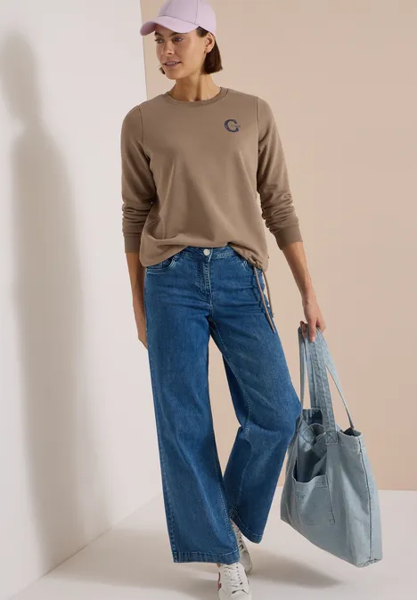 Frau in beigem Pullover, weiter Jeans, rosa Mütze und hellblauem Stoffbeutel vor hellem Hintergrund.