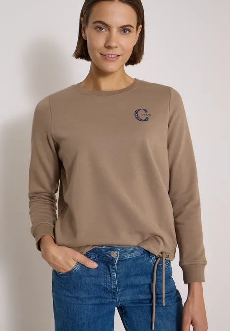 Frau trägt beigen Pullover mit kleinem Logo und blaue Jeans.