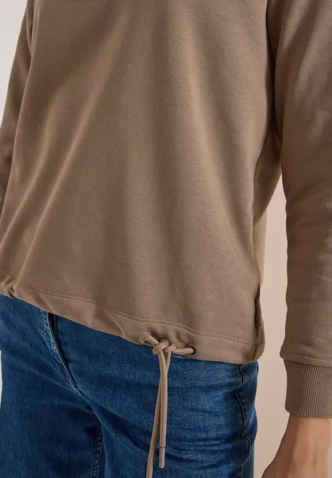 Braunes Langarmshirt mit Kordelzug am Saum, kombiniert mit blauen Jeans.