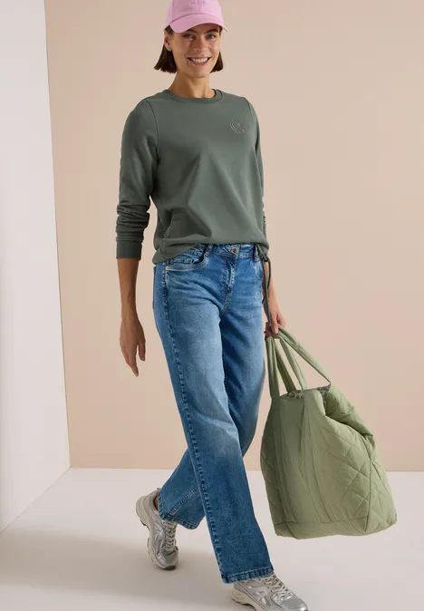 Frau mit grünem Pullover, Jeans, Sneakers, rosa Cap und großer grüner Tasche vor neutralem Hintergrund.