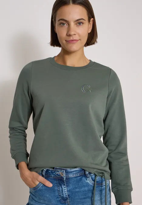 Frau trägt grünes Sweatshirt mit kleinem Logo und blaue Jeans vor hellem Hintergrund.