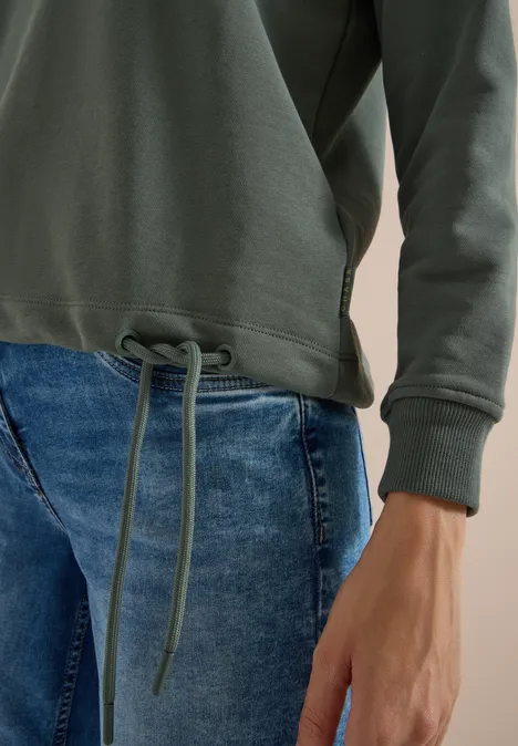 Person trägt olivgrünes Sweatshirt mit Tunnelzug am Saum und blaue Jeans, nur Oberkörper und Hand sichtbar.