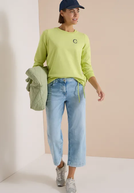 Frau in leuchtend grünem Langarmshirt, Jeans und silbernen Sneakers, hält passende Jacke in der Hand.